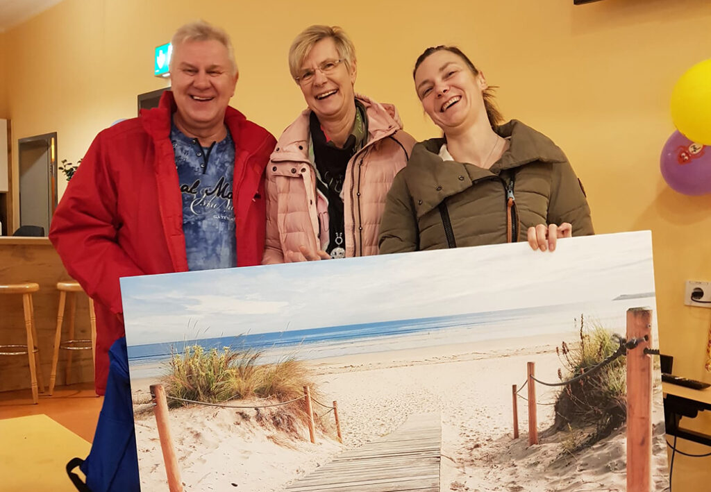 Drei Personen halten ein gro&szlig;es Landschaftsbild eines Strandes in einem Innenraum.