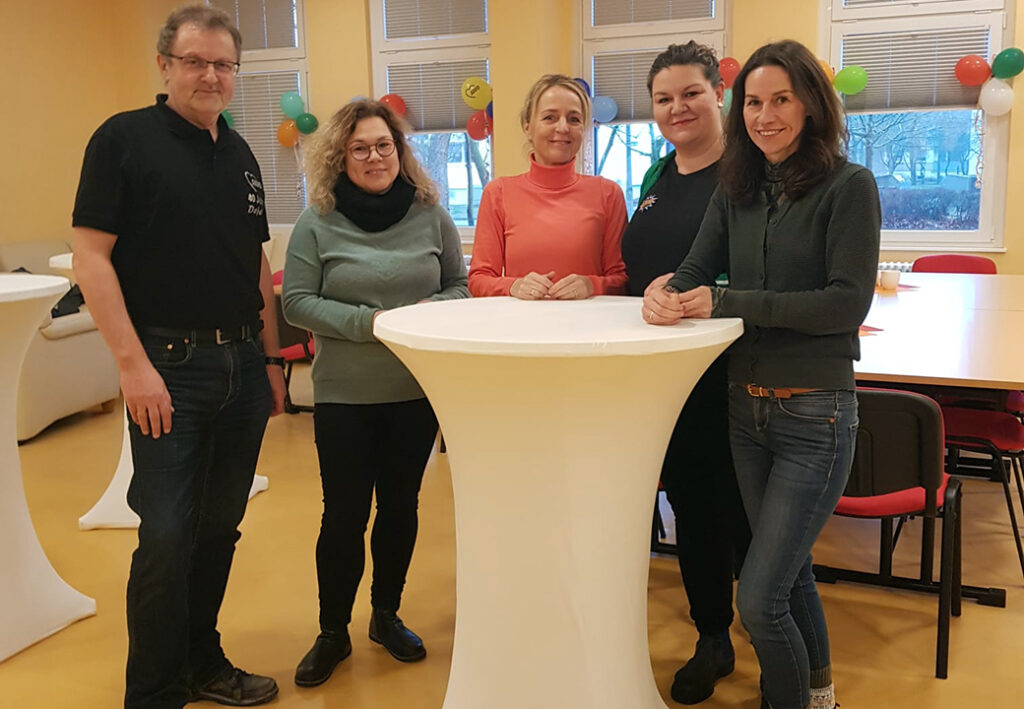 F&uuml;nf Personen stehen um einen wei&szlig;en Stehtisch in einem festlich dekorierten Raum.