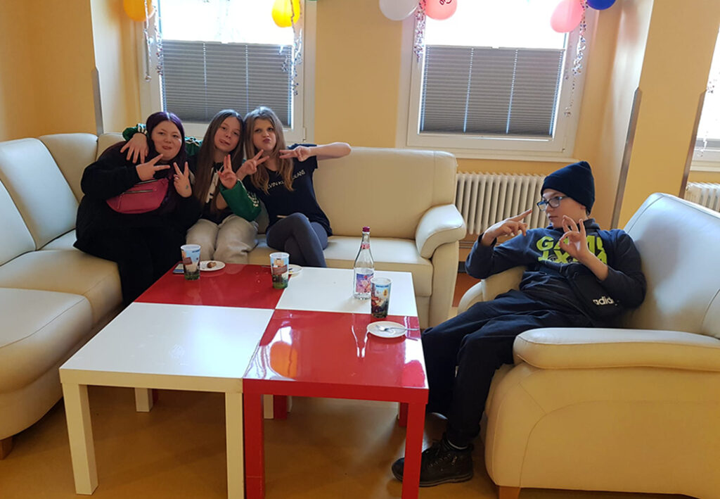 Vier Jugendliche sitzen auf Sofas und zeigen Handzeichen, auf einem Tisch stehen Getr&auml;nke und Kuchen.