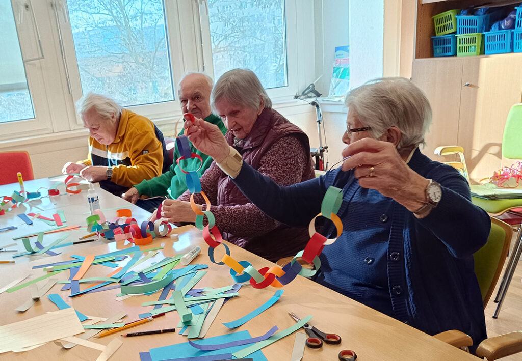 Eine Gruppe &auml;lterer Menschen bastelt farbige Papiergirlanden.