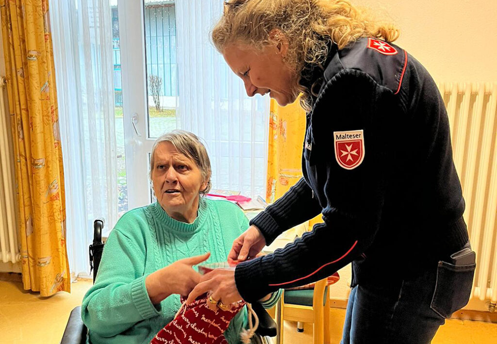 Eine Frau im Rollstuhl erhält ein Geschenk von einer Person in Malteser-Uniform.