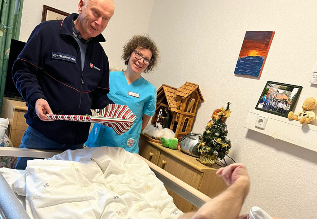 Ein Mann und eine Frau stehen l&auml;chelnd neben einem Krankenbett mit Geschenken.