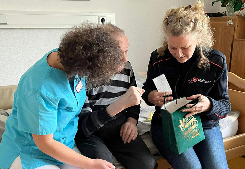 Zwei Frauen und ein &auml;lterer Mann sitzen auf einem Sofa, die Frauen helfen dem Mann dabei, eine kleine Geschenkt&uuml;te zu &ouml;ffnen.