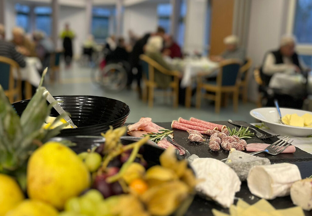 Tisch mit Fleisch und K&auml;se in einem Speisesaal mit Menschen im Hintergrund.