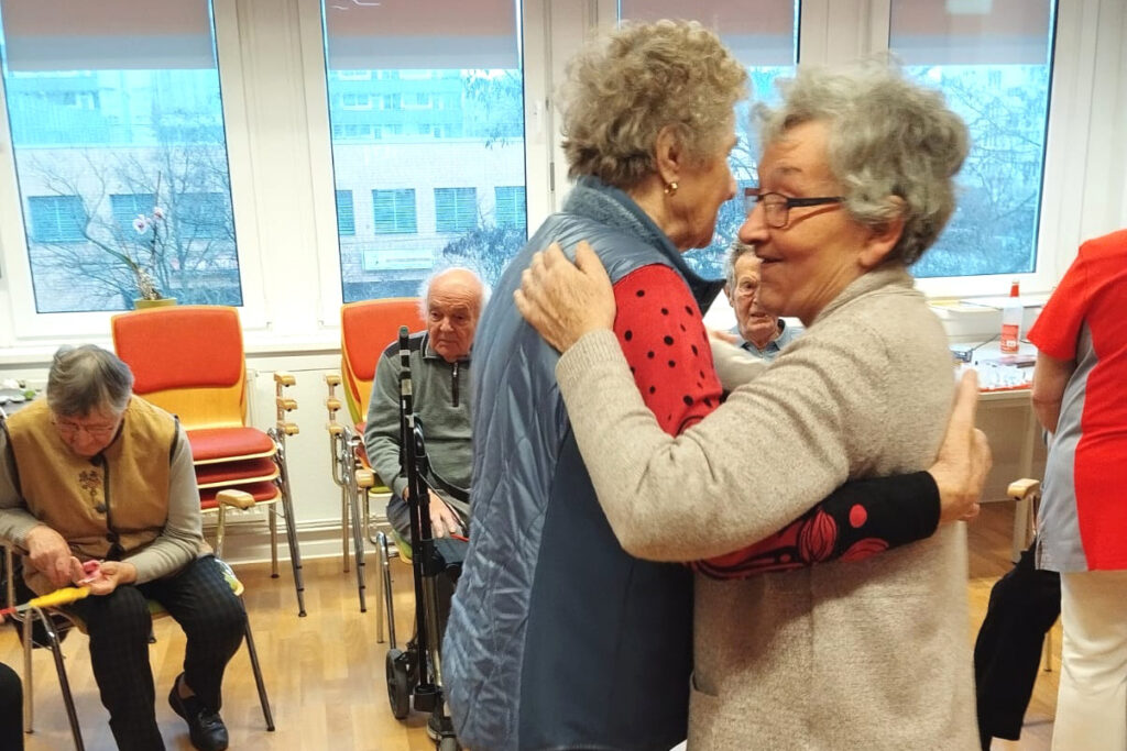 &Auml;ltere Menschen in einem Gemeinschaftsraum. Zwei Frauen umarmen sich, w&auml;hrend andere Personen auf St&uuml;hlen sitzen und zuschauen.