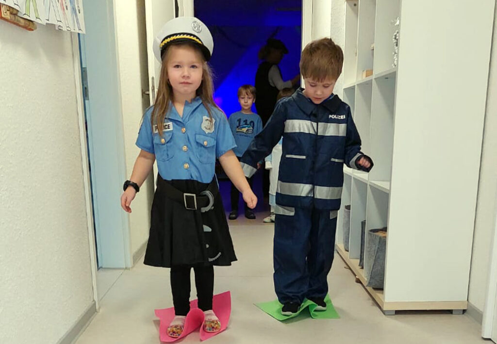 Kinder in Polizeiuniformen halten sich an den H&auml;nden und gehen auf farbigen Bl&auml;ttern einen Flur entlang.