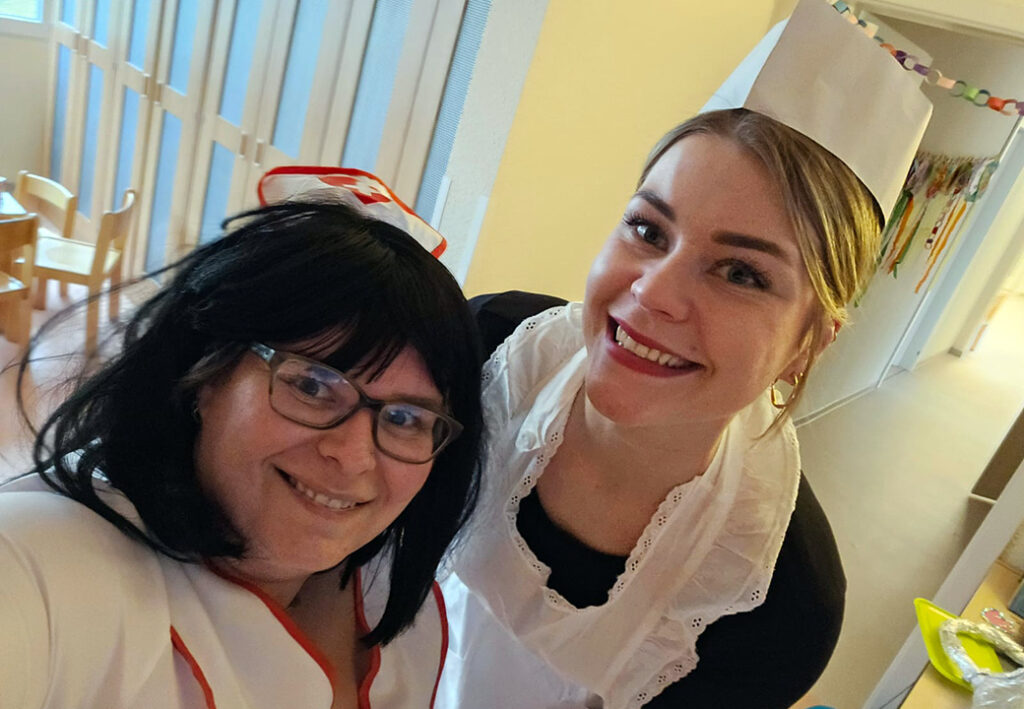 Zwei Personen in Verkleidung, eine mit Krankenschwester-Kost&uuml;m und die andere mit einer Art Dienstm&auml;dchen-Outfit, l&auml;cheln in die Kamera.