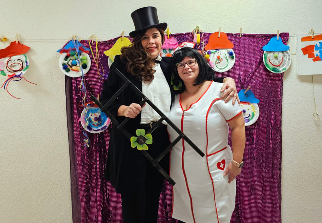 Zwei verkleidete Personen posieren lachend vor einer bunten Dekoration.