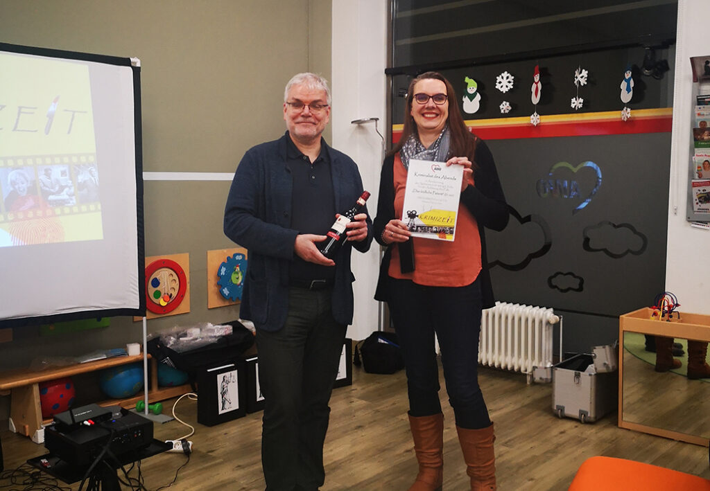 Zwei Personen stehen in einem Raum, eine h&auml;lt eine Urkunde und eine Flasche Wein, die andere eine Urkunde mit Aufdruck 'Krimizeit'.