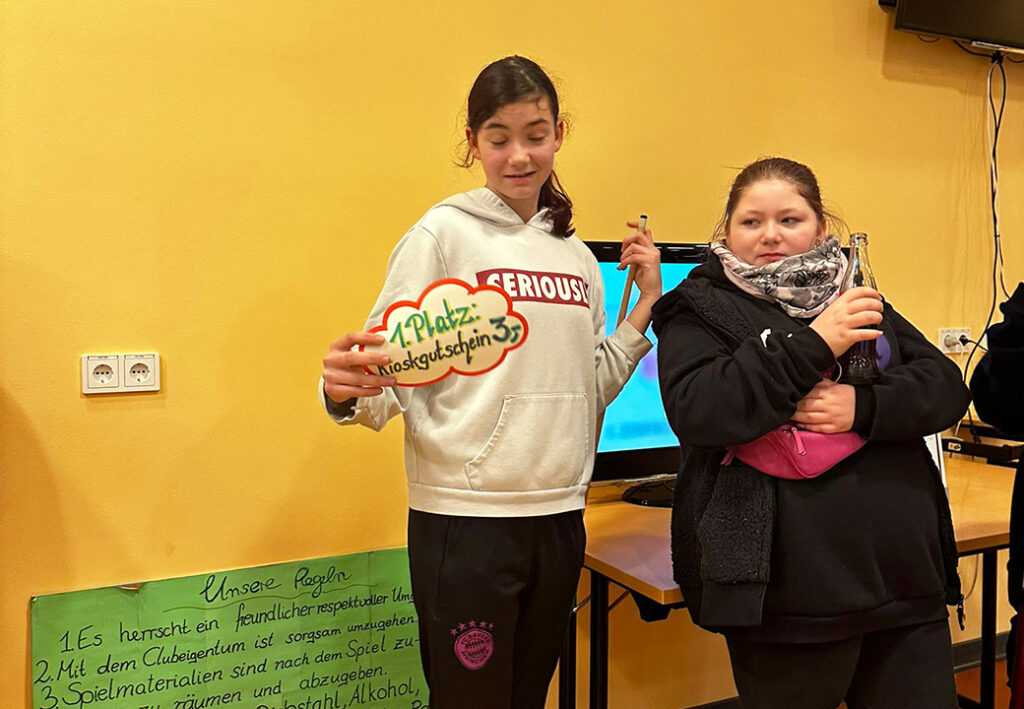 Zwei Jugendliche in einem Raum, eine hält ein Schild mit '1. Platz: Kioskgutschein'