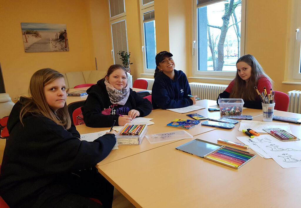 Vier Jugendliche sind in einem Raum an einem Tisch beim Malen und Zeichnen. Auf dem Tisch liegen Buntstifte und Papier.