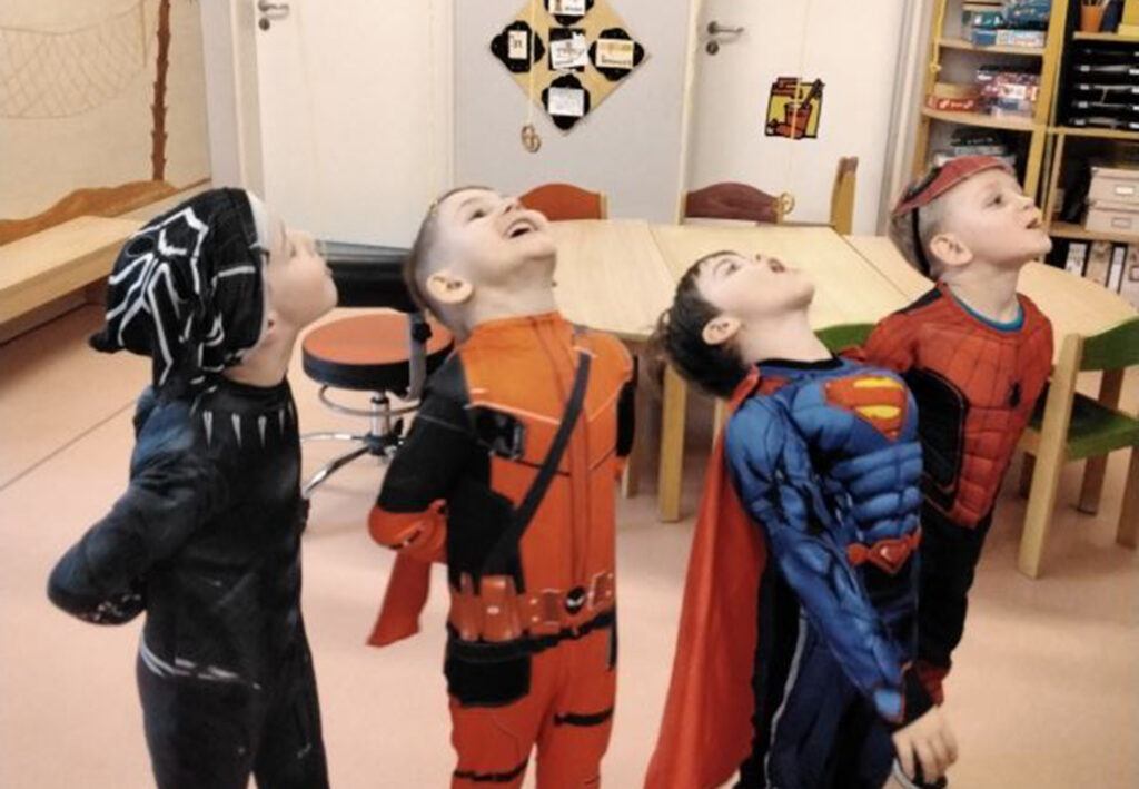 Vier Kinder in Superheldenkost&uuml;men stehen nebeneinander in einem Raum und schauen nach oben.