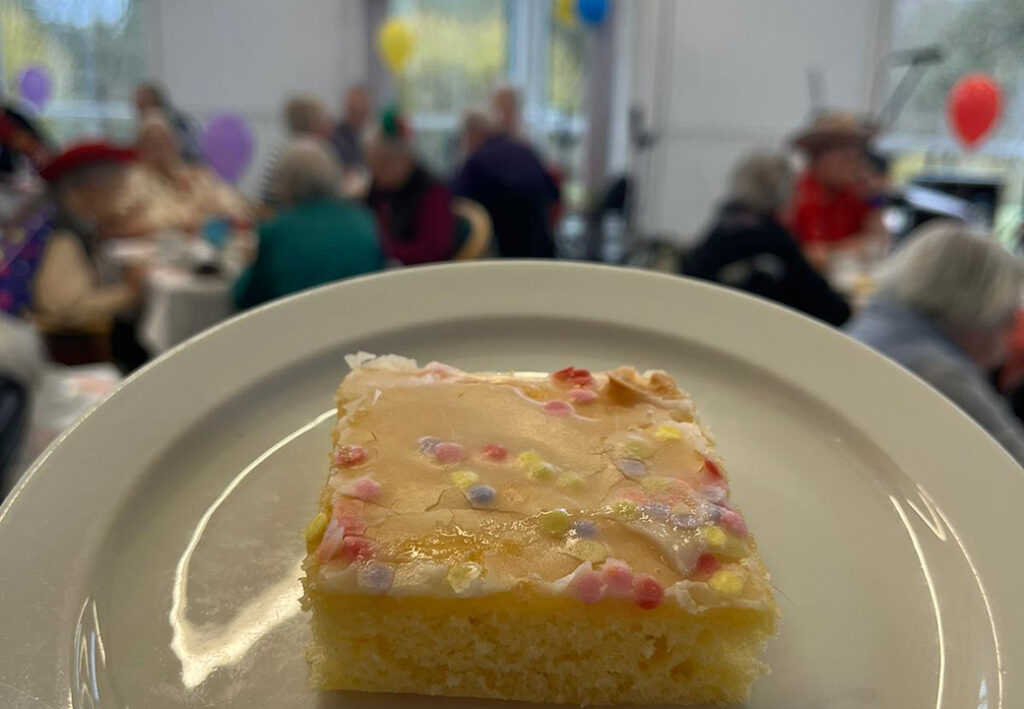 Ein St&uuml;ck Kuchen auf einem Teller im Vordergrund, mit unscharfem Hintergrund einer Gruppenveranstaltung.