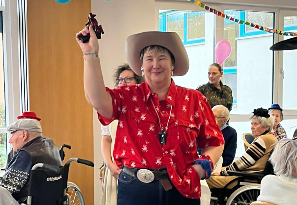 Person in rotem Hemd und Cowboyhut h&auml;lt ein Spielzeug in der Hand, umgeben von Menschen, einige im Rollstuhl.