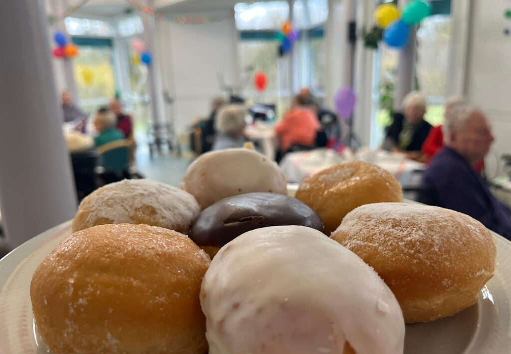 Ein Teller mit Berliner Pfannkuchen steht im Vordergrund eines Raumes mit Menschen an Tischen, dekoriert mit bunten Luftballons.