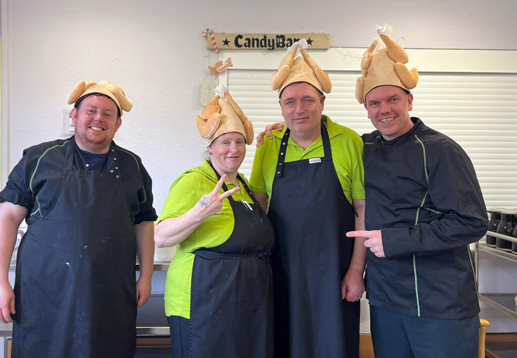 Vier Personen in Kochsch&uuml;rzen und H&uuml;ten in Form von Truth&auml;hnen posieren l&auml;chelnd f&uuml;r ein Foto.