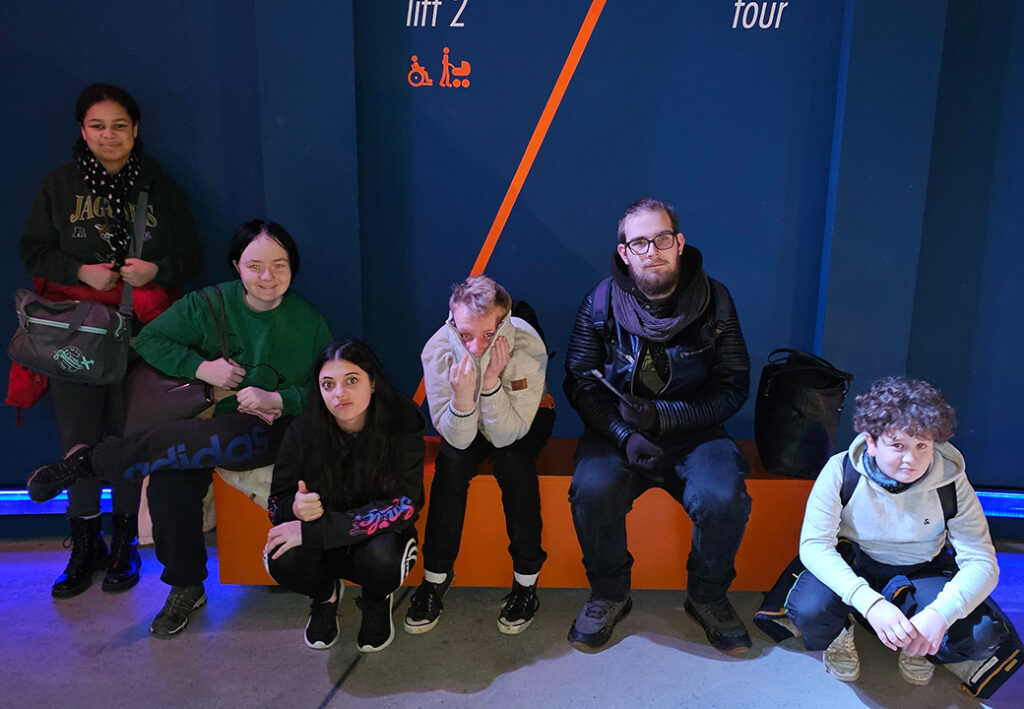Eine Gruppe von sechs Personen sitzt und steht vor einem blauen Hintergrund in einem Indoor-Bereich.