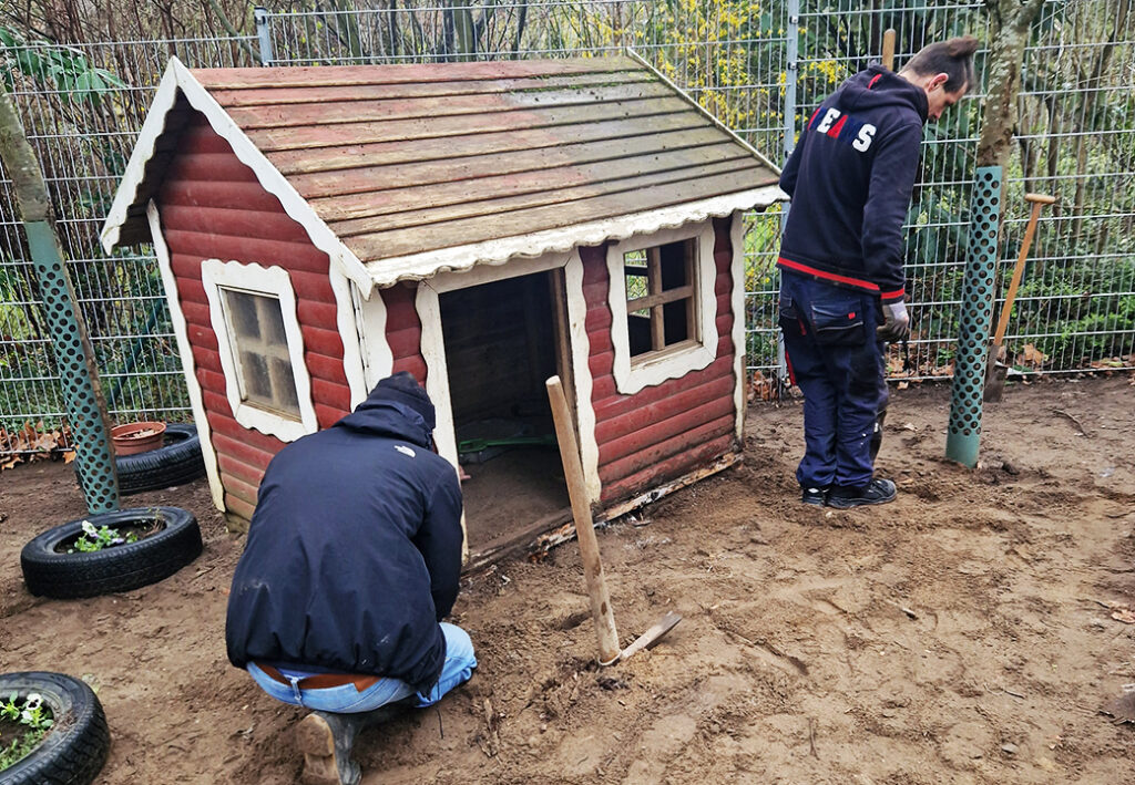 Zwei Personen, die im Freien vor einem kleinen Holzh&auml;uschen arbeiten.