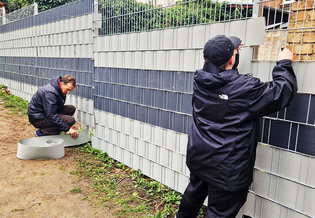 Zwei Personen befestigen Lamellen an einem Zaun.