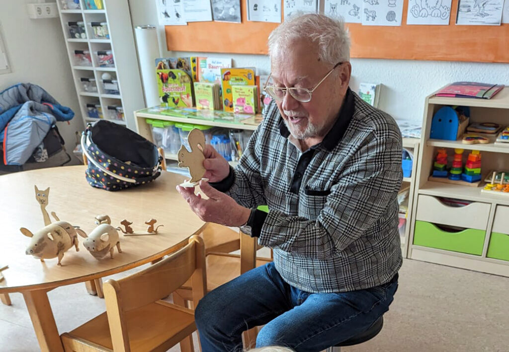 Ein &auml;lterer Mann, der in einem Raum mit Kinderspielzeug sitzt und ein Holzspielzeug in Form eines Tiers in den H&auml;nden h&auml;lt.