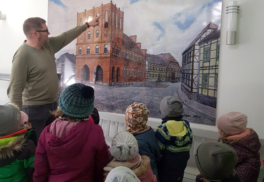 Ein Erwachsener zeigt einer Gruppe von Kindern ein gro&szlig;es Bild oder Gem&auml;lde eines historischen Geb&auml;udes.