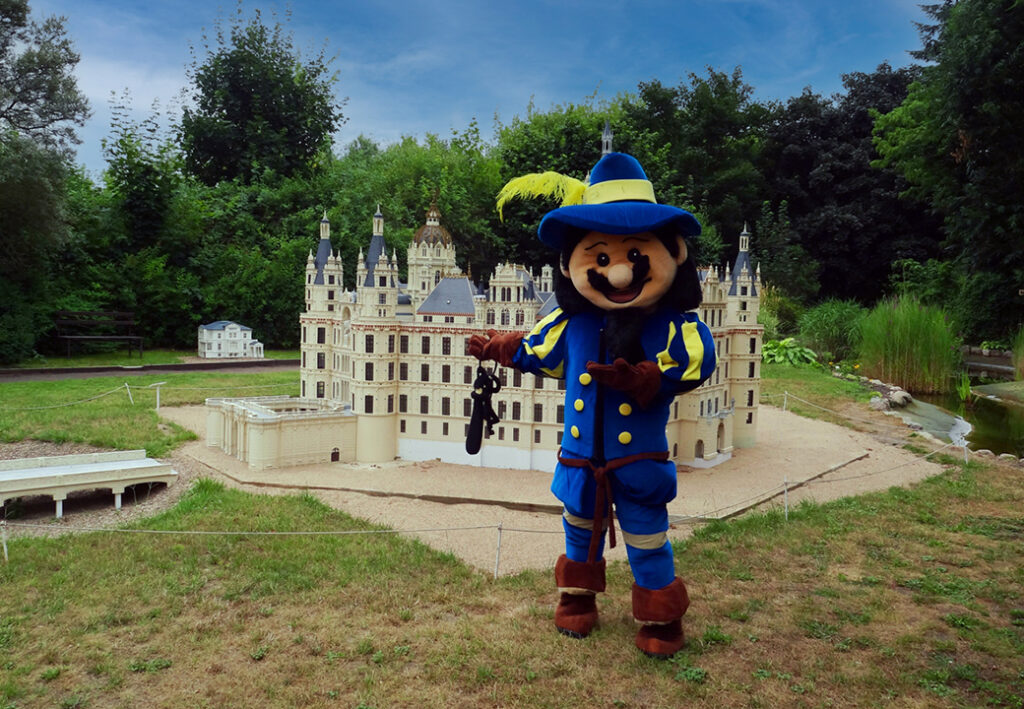 Mensch in einem farbenfrohen Maskottchenkostüm steht vor einem Modell eines Schlosses im Freien.