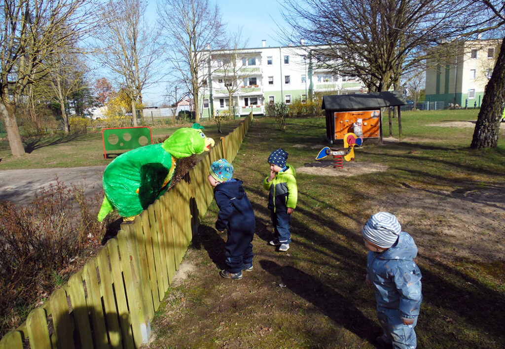 Kinder auf einem Spielplatz beobachten ein gro&szlig;es gr&uuml;nes Pl&uuml;schtier &uuml;ber einem Zaun.