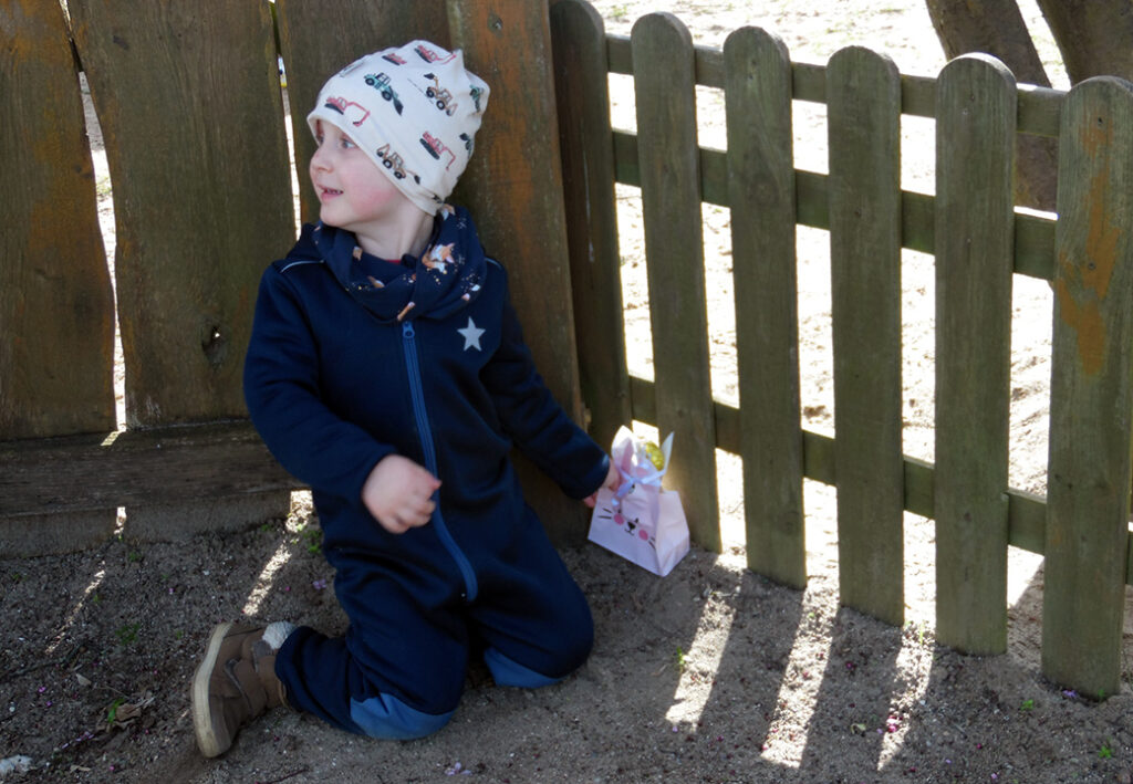 Kleinkind im blauen Anzug mit M&uuml;tze und Osterk&ouml;rbchen kniet im Sand vor einem Zaun.