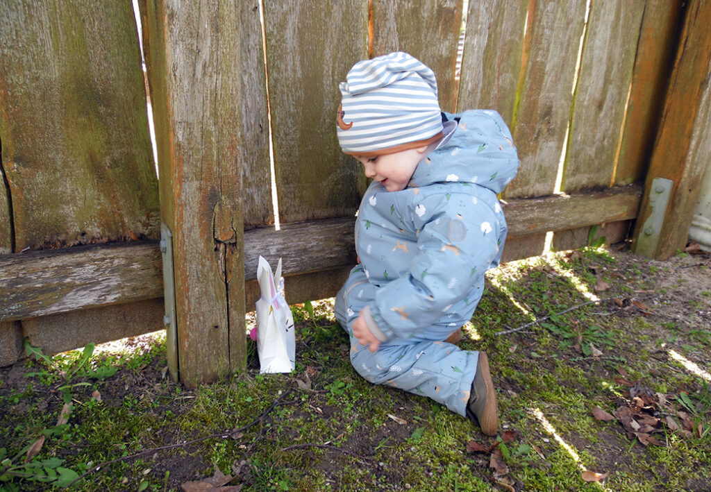 Ein Kleinkind in einem blauen Schneeanzug sitzt auf dem Boden vor einem Holzzaun und betrachtet eine kleine T&uuml;te mit Hasen-Motiv.