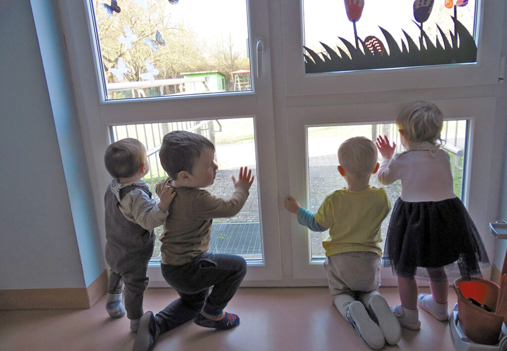Vier kleine Kinder blicken aus einer Glast&uuml;r nach drau&szlig;en.