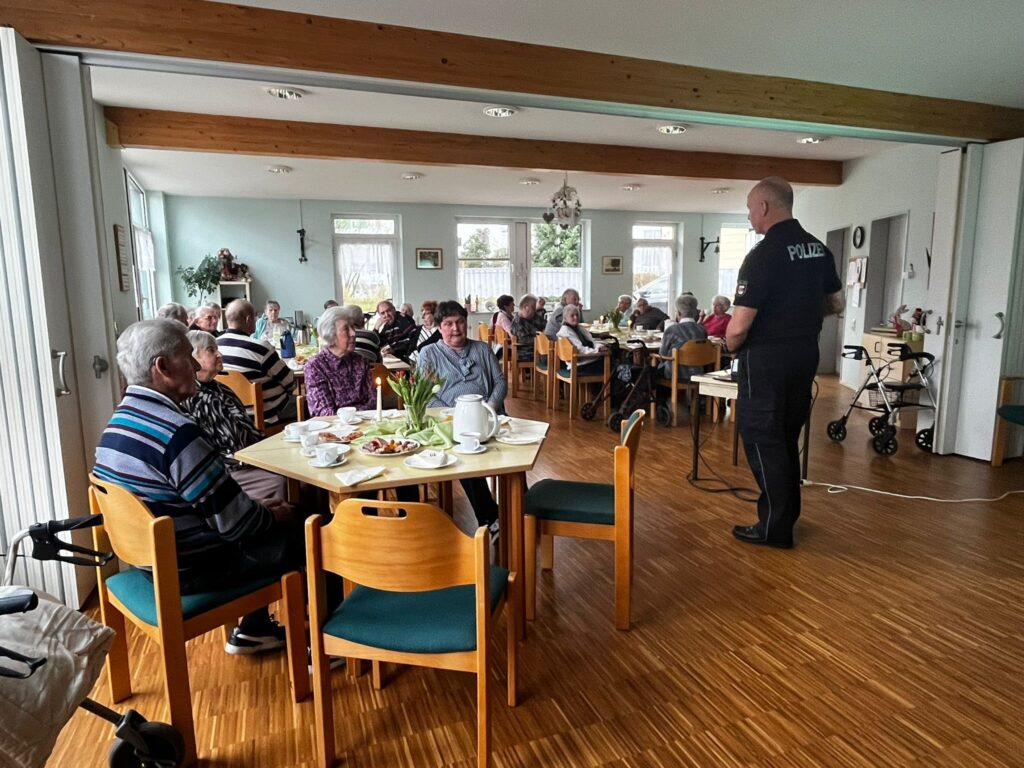 Gruppe von älteren Menschen in einem Gemeinschaftsraum, an verschiedenen Tischen sitzend und einem Polizisten zuhörend.