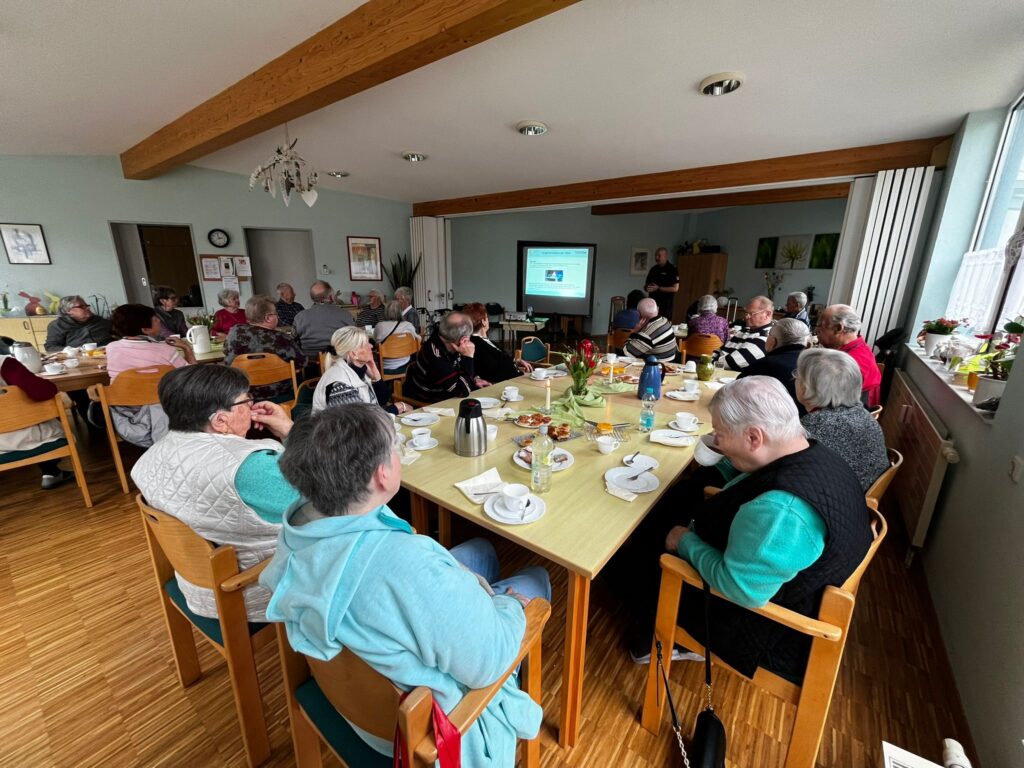 Eine Gruppe &auml;lterer Menschen in einem Gemeinschaftsraum, die an Tischen sitzen und einer Pr&auml;sentation folgen.