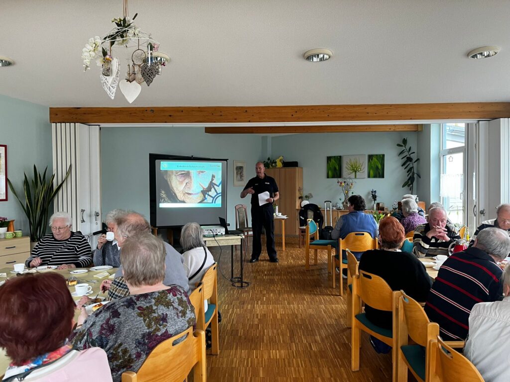 Eine Gruppe &auml;lterer Menschen sitzt in einem Gemeinschaftsraum an Tischen und h&ouml;rt einem Vortrag zu.