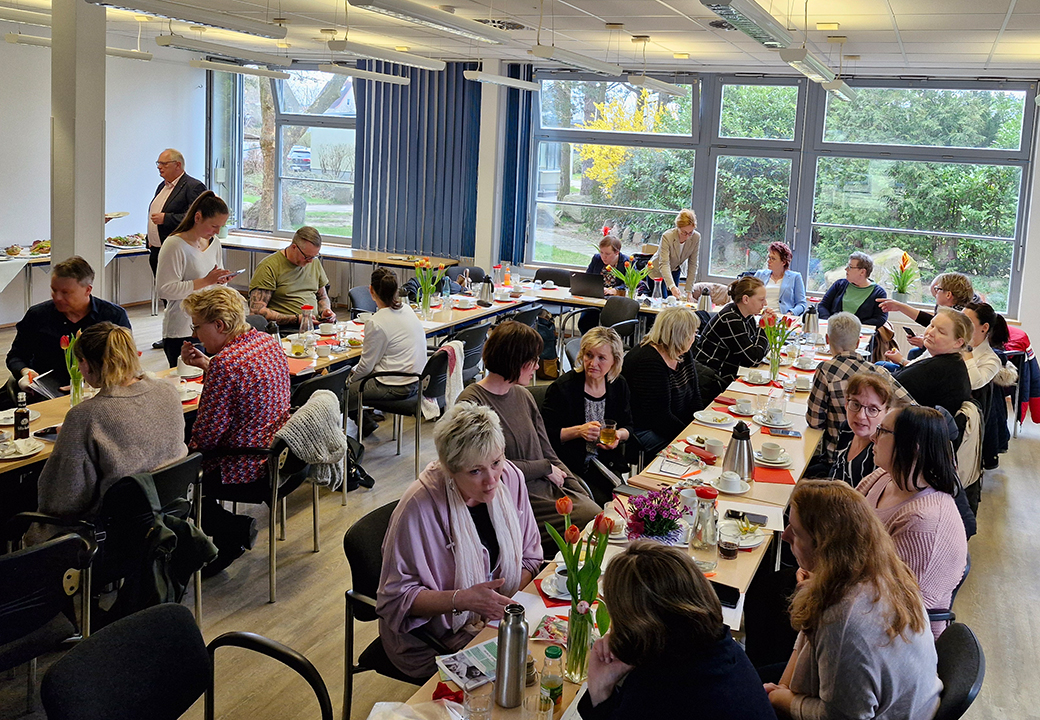 Menschen sitzen an langen Tischen in einem hellen Raum.