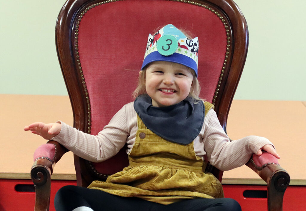 Ein Kind mit einer Krone sitzt auf einem roten Stuhl.