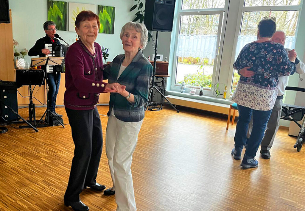 Zwei &auml;ltere Frauen tanzen gemeinsam in einem hellen Raum, im Hintergrund spielt ein Musiker Akkordeon.