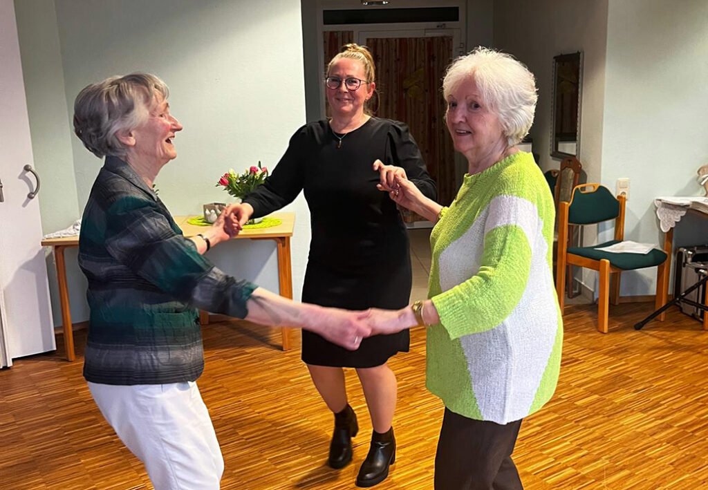 Drei Frauen tanzen Hand in Hand in einem Raum mit Holzboden.