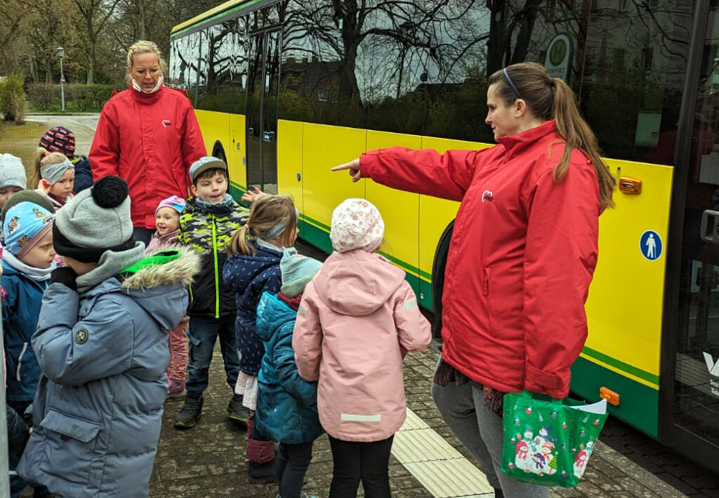 Zwei Frauen in roten Jacken, die eine Gruppe Kinder in Winterkleidung vor einem gelben Bus anleiten.