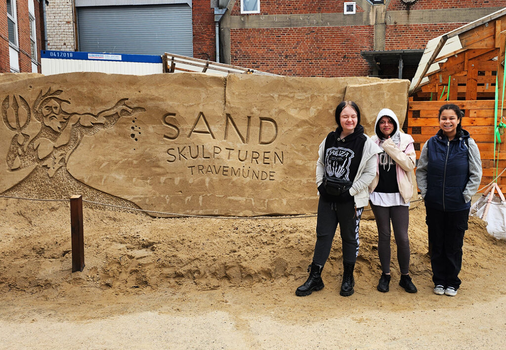 Drei Personen stehen vor einer Sandskulptur mit einer Inschrift.