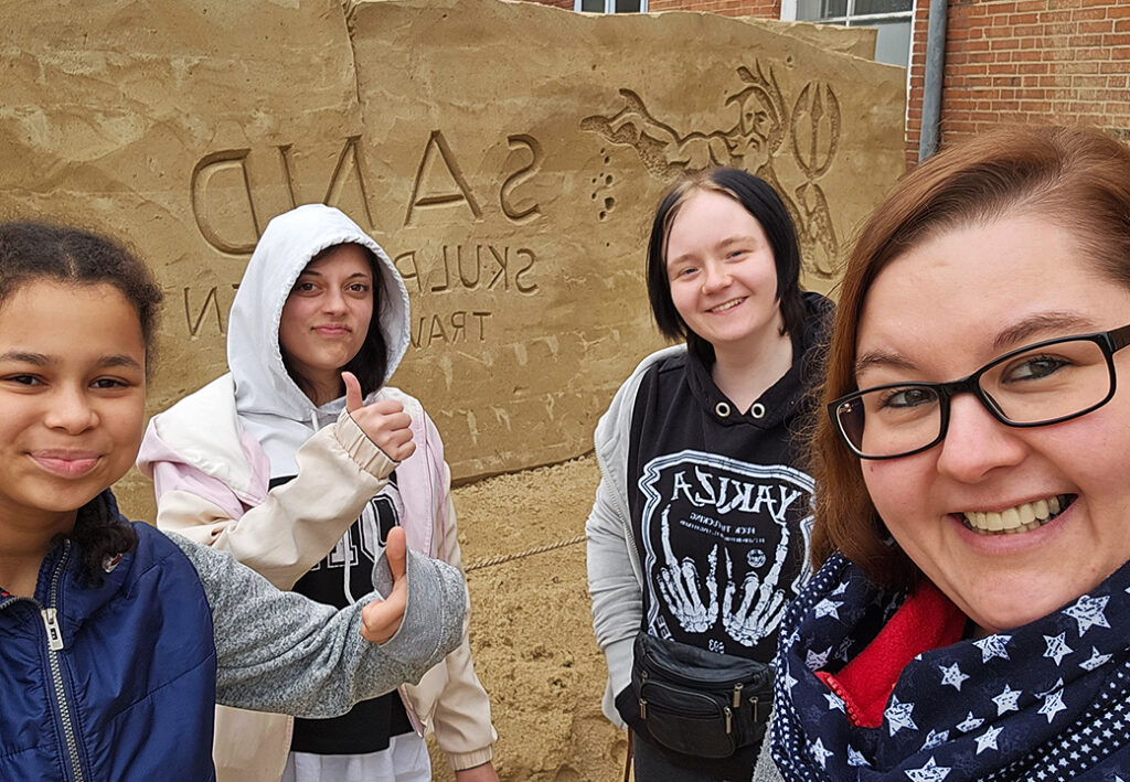 Vier Personen l&auml;cheln vor einer Sandskulptur in die Kamera.