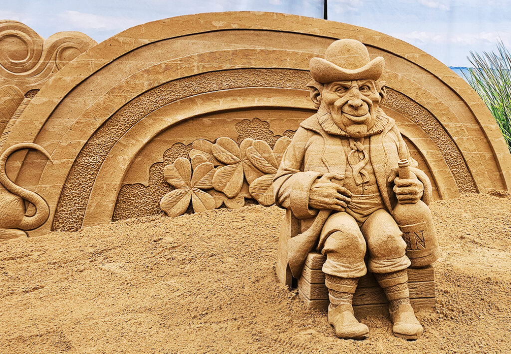 Eine Sandskulptur eines sitzenden Mannes mit einem Hut und einer Flasche in der Hand.