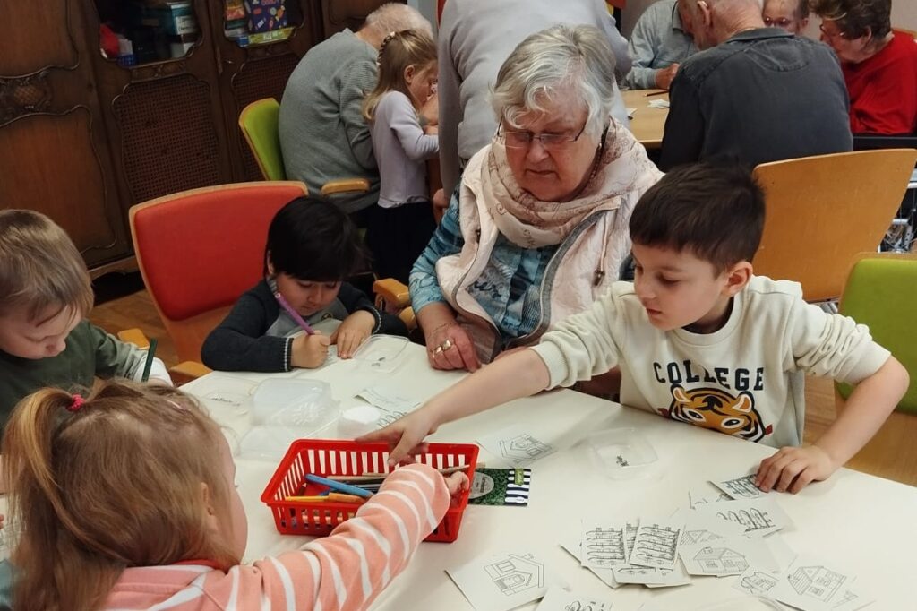 Mehrere Kinder und eine &auml;ltere Frau sitzen an einem Tisch und basteln gemeinsam mit Papier und Stiften.