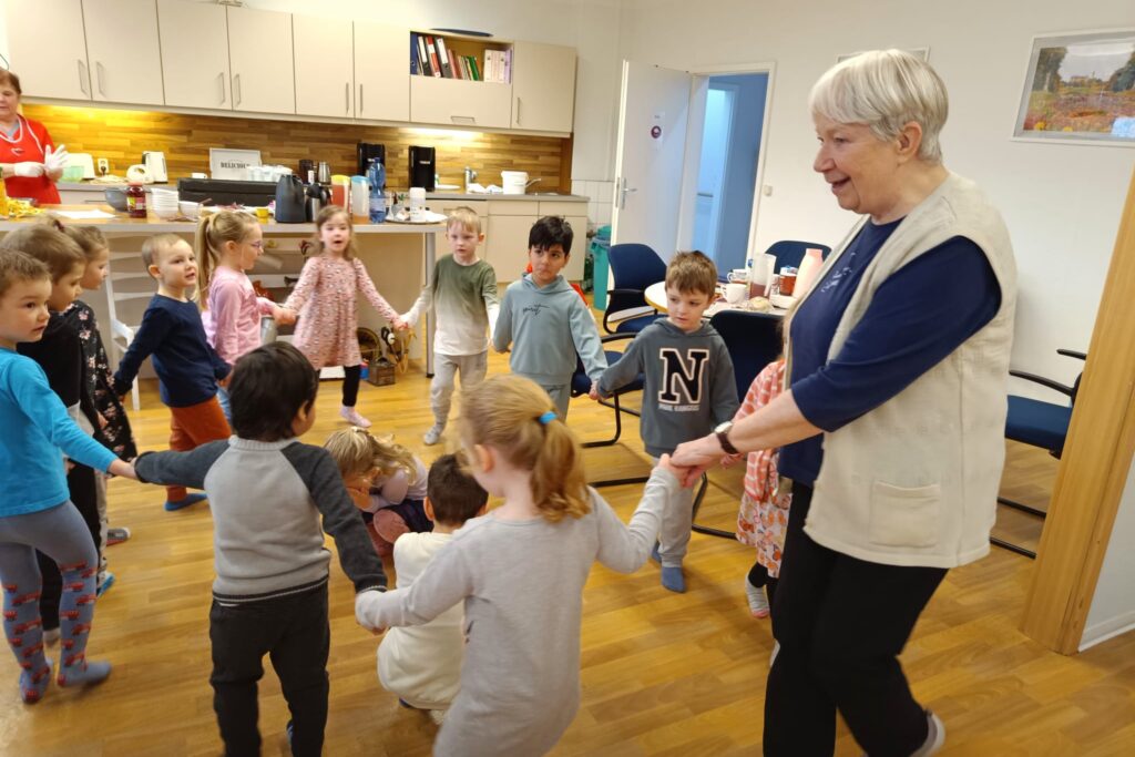 Eine Gruppe von Kindern spielt im Kreis mit einer älteren Frau in einem Raum mit Küchenzeile.