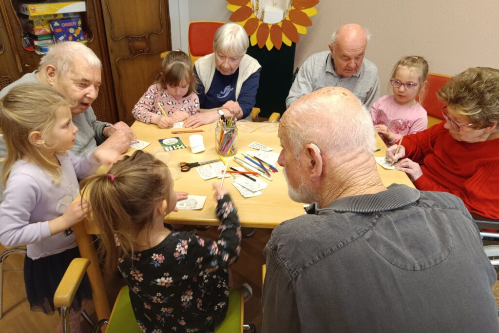 &Auml;ltere Menschen und Kinder sitzen an einem Tisch und malen zusammen.