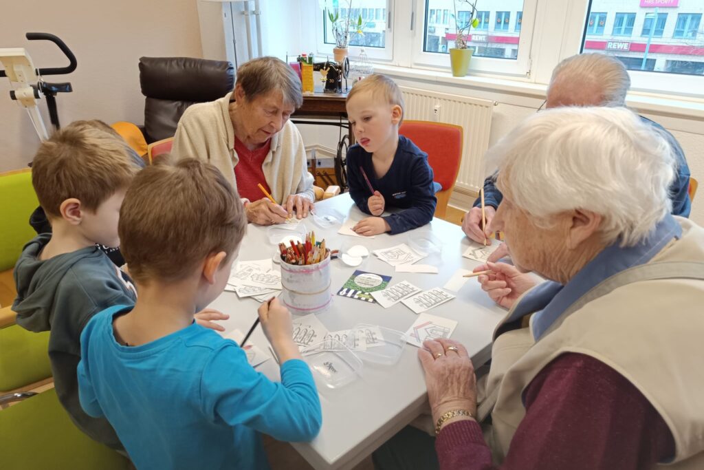 &Auml;ltere und j&uuml;ngere Menschen sitzen zusammen am Tisch und malen.