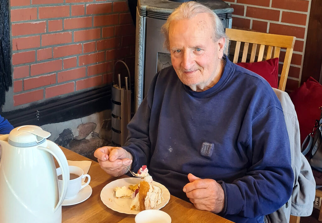 Ein &auml;lterer Mann sitzt an einem Tisch in einem Ziegelsteinraum und genie&szlig;t ein St&uuml;ck Torte neben einer Kaffeekanne.