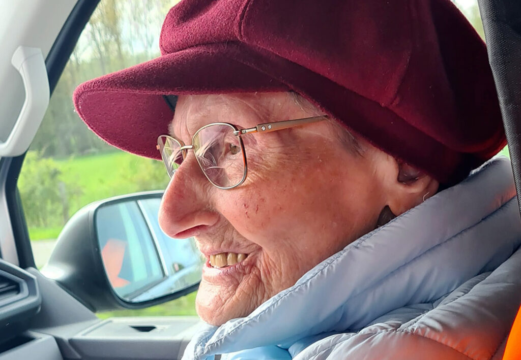 &Auml;ltere Frau mit Brille und rotem Hut sitzt in einem Auto und l&auml;chelt.