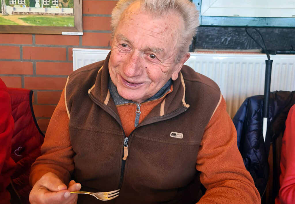 Ein &auml;lterer Mann in brauner Fleecejacke sitzt l&auml;chelnd an einem Tisch und h&auml;lt eine Gabel in der Hand.