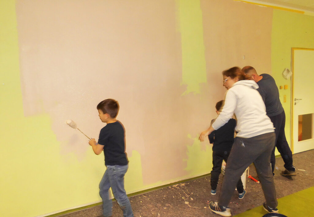Eine Gruppe von Menschen, darunter zwei Kinder und zwei Erwachsene, streichen gemeinsam eine Wand in einem Raum.