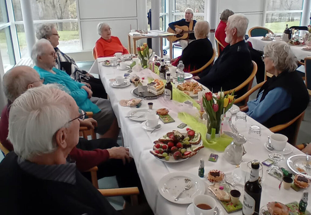 &Auml;ltere Menschen sitzen an einem festlichen Tisch und h&ouml;ren einer Person zu, die Gitarre spielt.